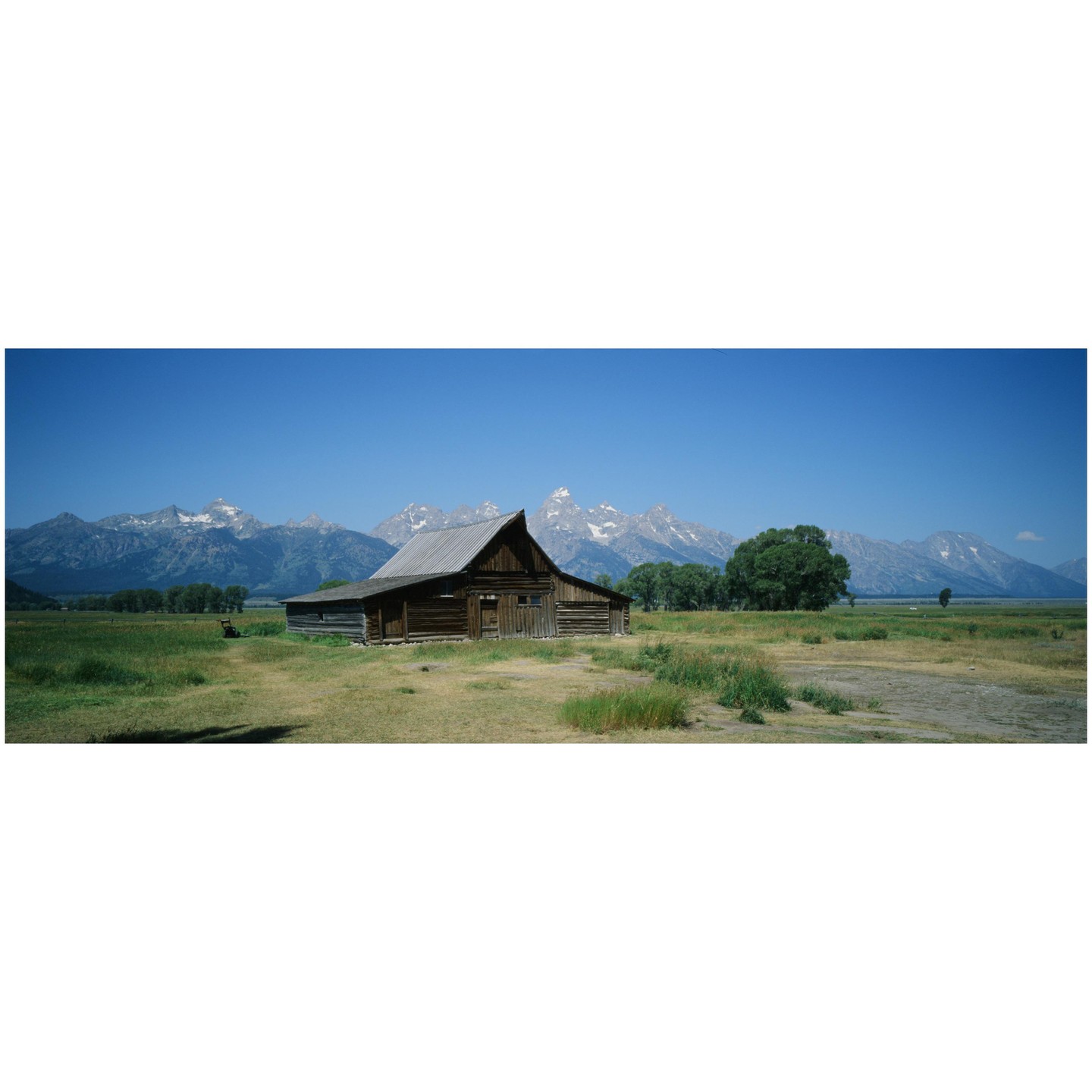 A wooden barn stands in a field with snow-capped mountains in the background under a clear blue sky.