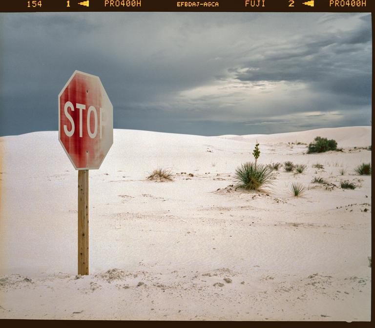 A stop sign stands in a desert landscape with white sand dunes and sparse vegetation under a cloudy sky.
