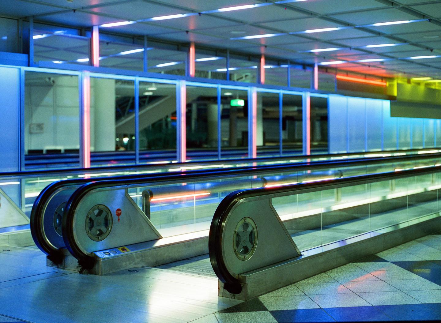 Two moving walkways are illuminated by colorful lights in an airport terminal at night.