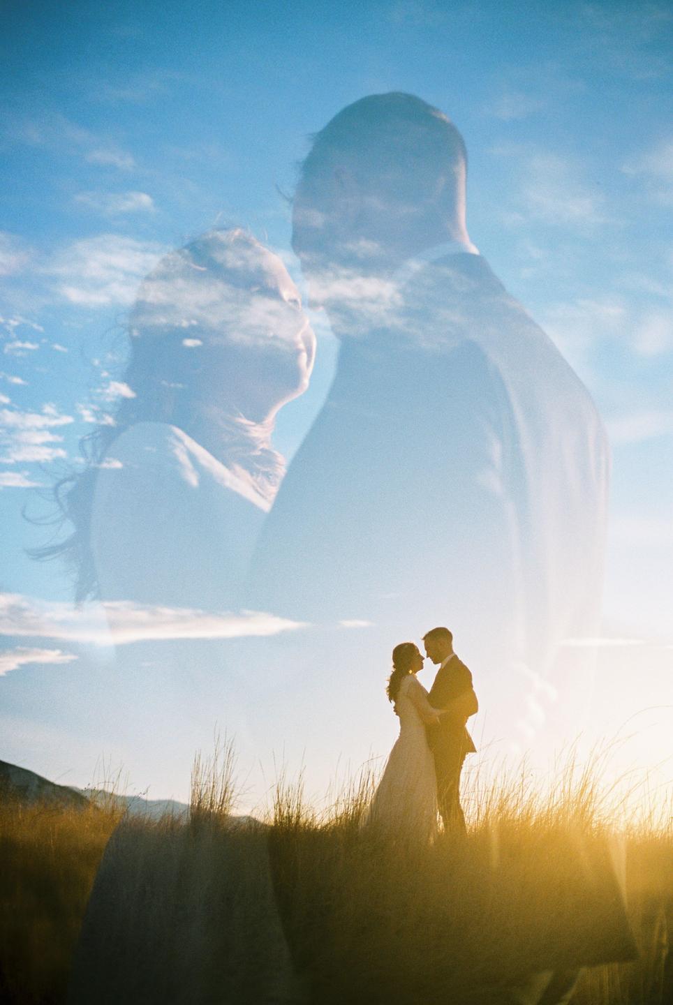 A couple stands in a field of tall grass at sunset, with a double exposure showing their faces against a cloudy sky.