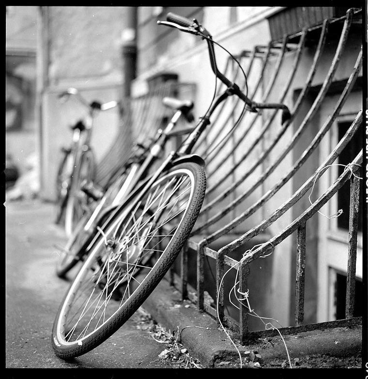 A bicycle leans against a metal grate next to a building, with another bicycle visible in the background.