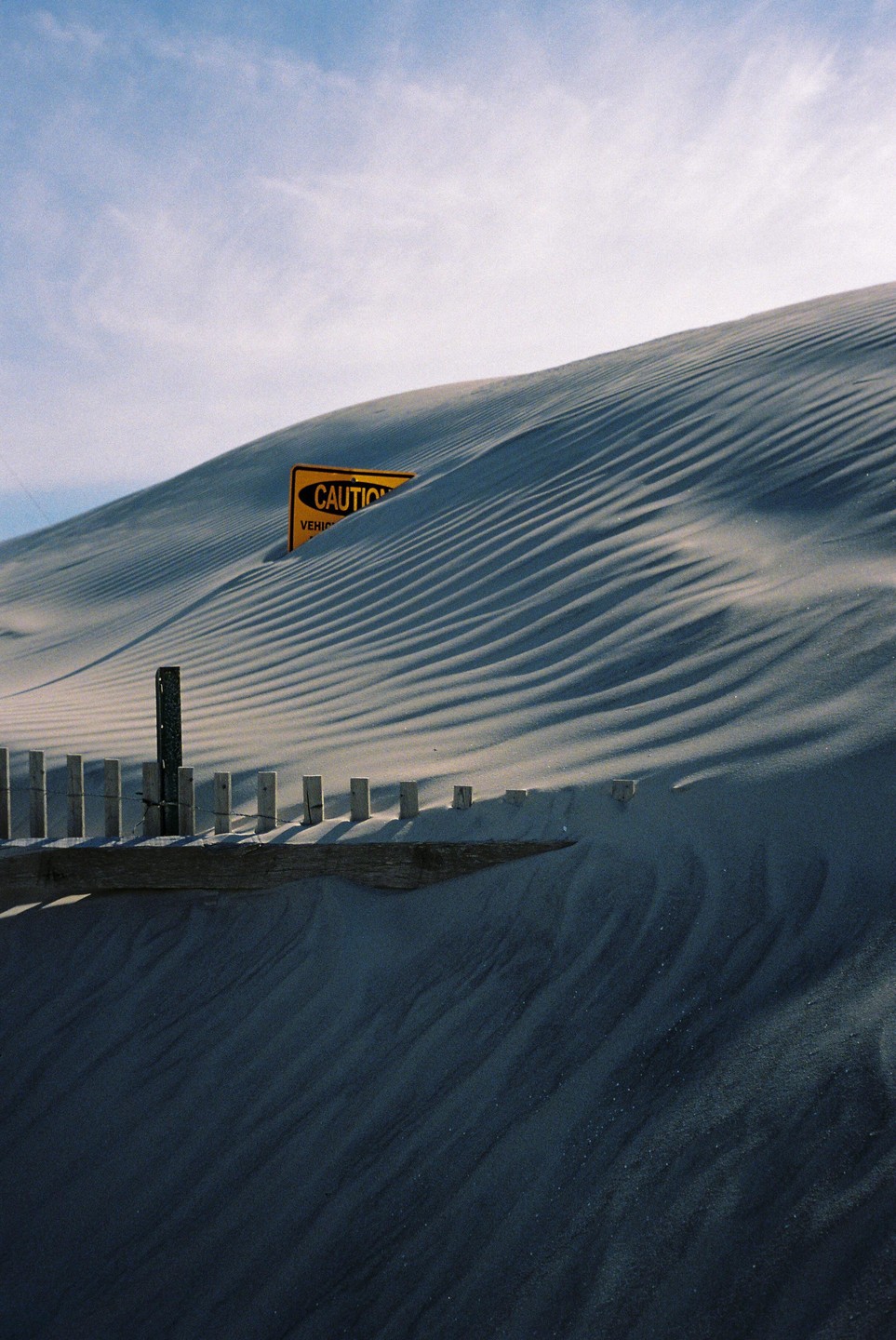 A yellow caution sign is partially buried in a sand dune with a wooden fence in the foreground.