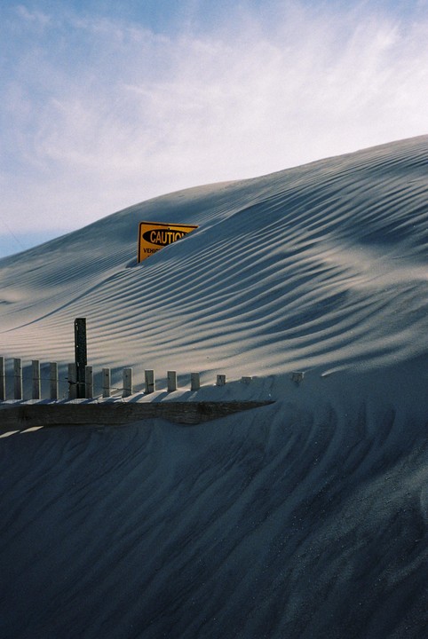 A yellow caution sign is partially buried in a sand dune with a wooden fence in the foreground.