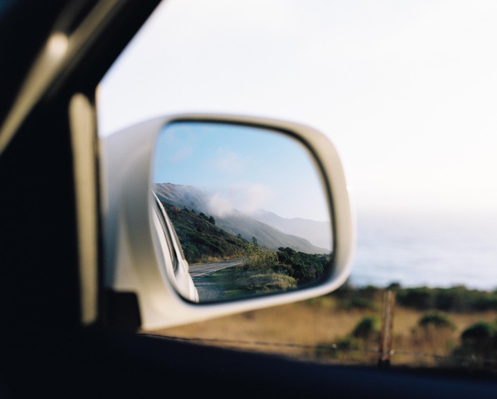 A car's side mirror reflects a winding road along a mountainous coast with fog.