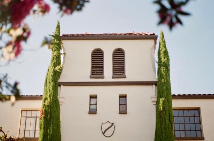 A cream-colored building with a tiled roof and two tall, narrow trees in front of it is shown under a clear sky.