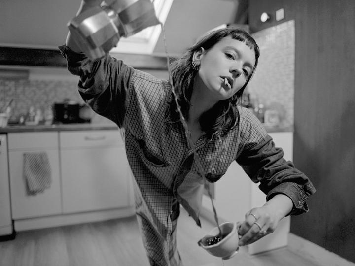 A woman with a cigarette in her mouth pours coffee from a moka pot into a cup in a kitchen.