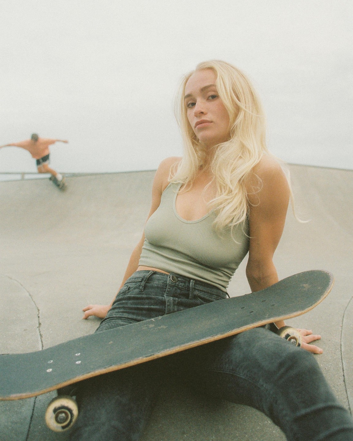 A woman sits in a skate park with a skateboard on her lap while a man skates in the background.
