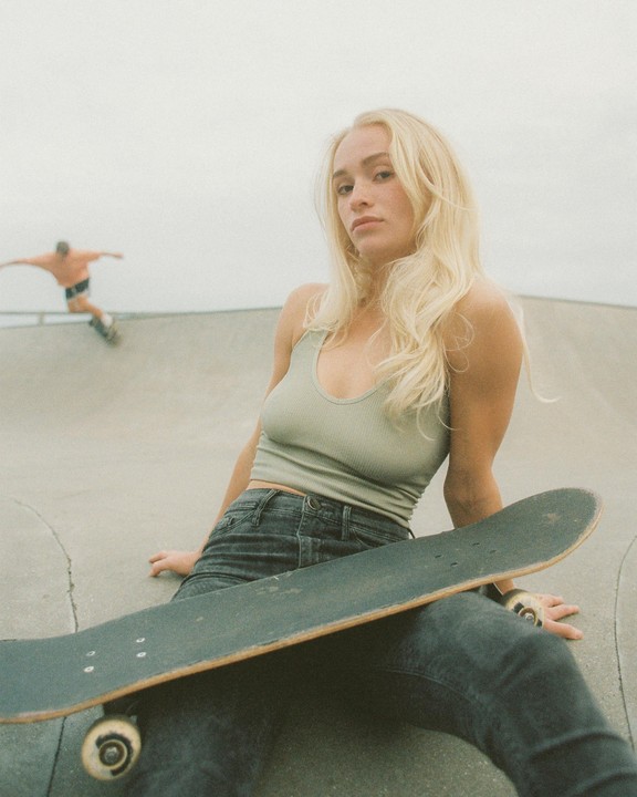 A woman sits in a skate park with a skateboard on her lap while a man skates in the background.