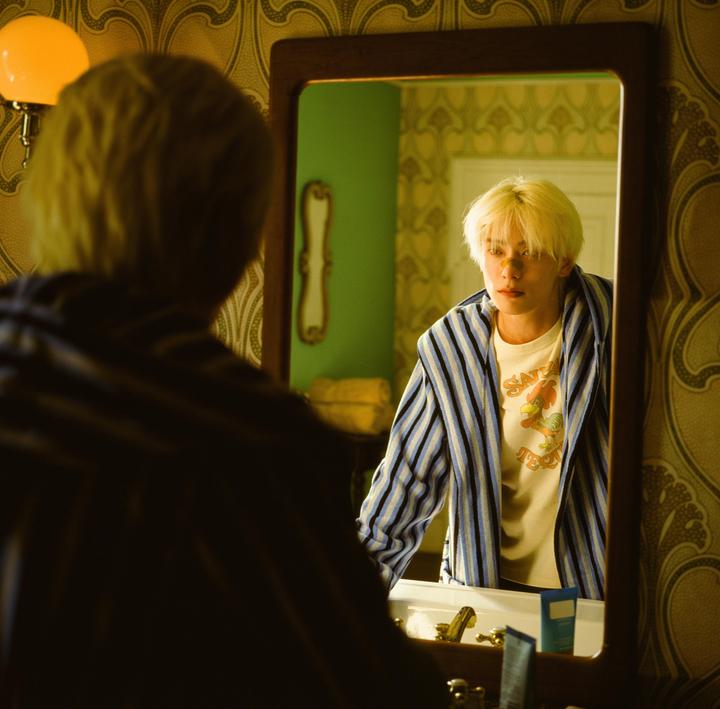 A man with blonde hair looks at his reflection in a mirror in a room with patterned wallpaper.