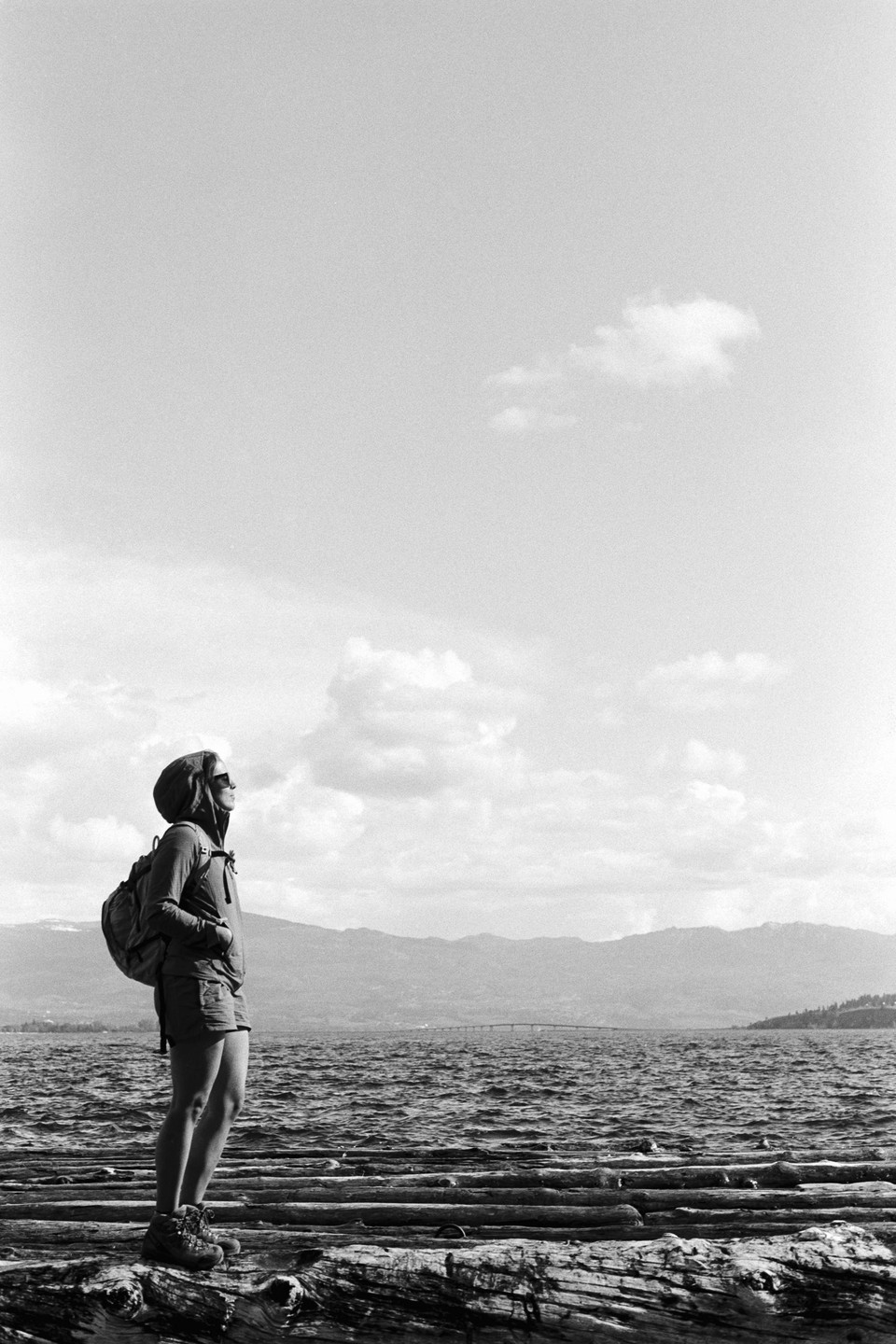 A woman wearing a hooded jacket, shorts, and a backpack stands on logs by a lake, looking up at the sky with mountains and a bridge in the distance.