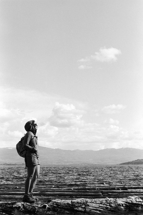 A woman wearing a hooded jacket, shorts, and a backpack stands on logs by a lake, looking up at the sky with mountains and a bridge in the distance.