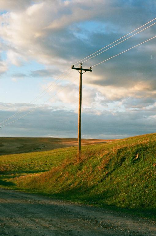 A telephone pole stands beside a dirt road and a grassy hill under a cloudy sky.