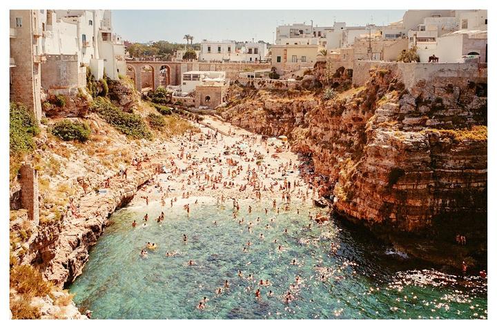 A crowded beach is nestled between rocky cliffs and white buildings under a clear sky.