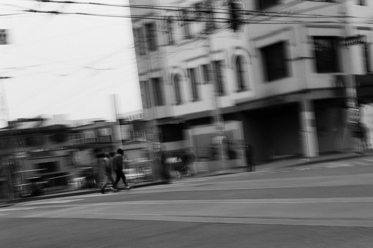 Two people walk across a street in front of a building with motion blur.
