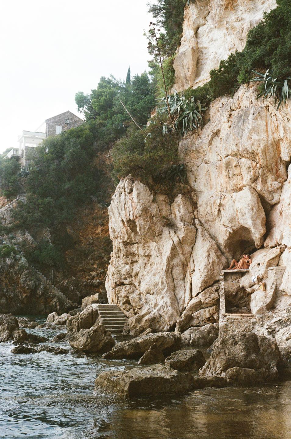A couple relaxes on a stone ledge by the sea, nestled against a rocky cliff with lush vegetation and a distant house.