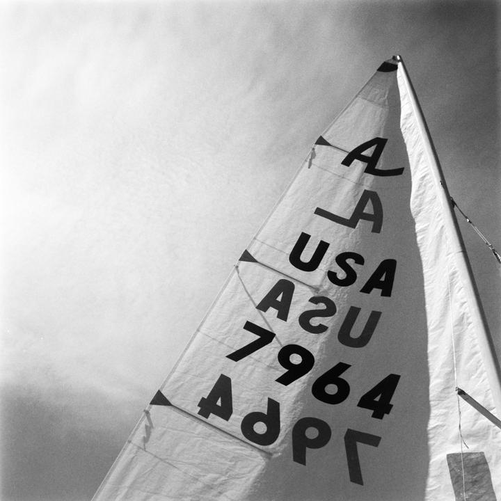A sailboat sail with "USA" and "1964" printed on it is angled against a bright sky.