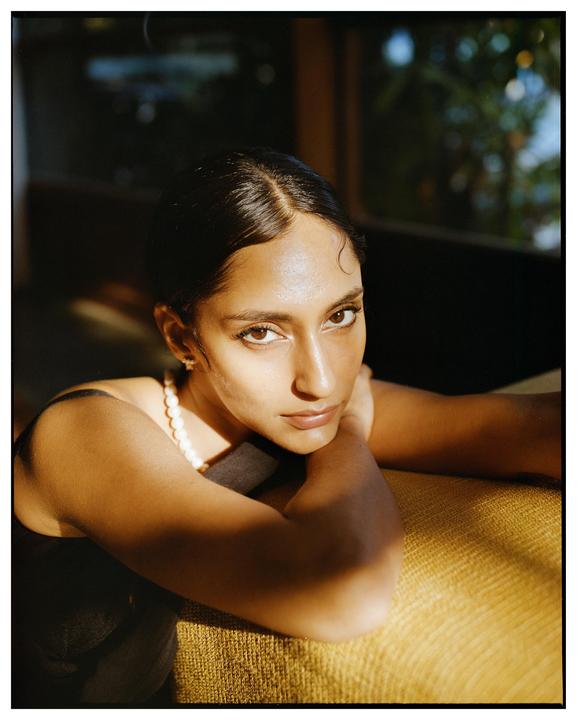 A woman with dark hair and a pearl necklace rests her arms on a textured surface, looking directly at the viewer.