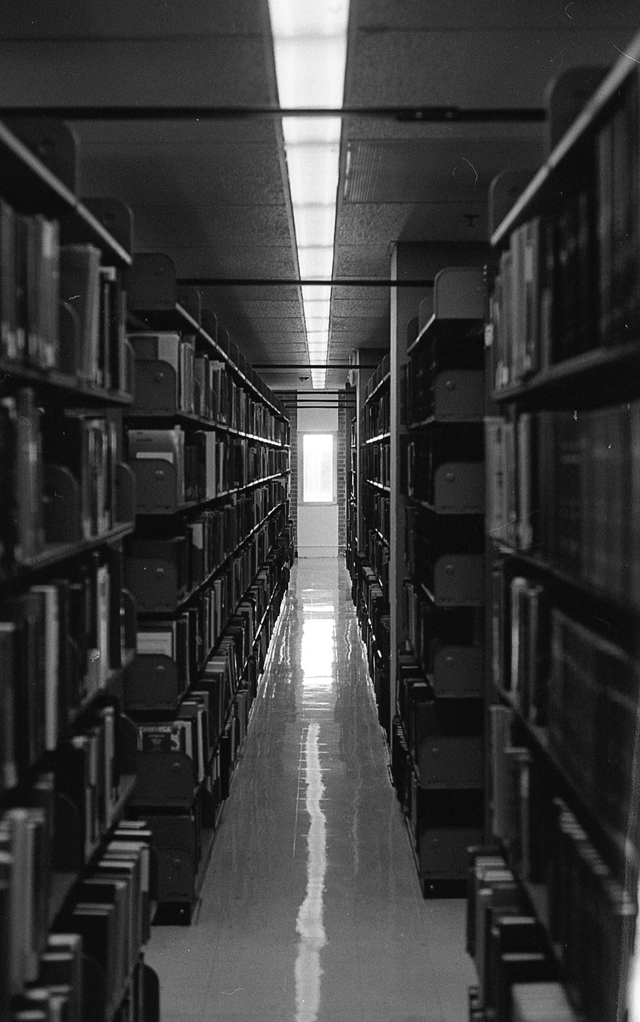 A long, narrow aisle in a library is lined with bookshelves on both sides, leading to a bright window at the far end.