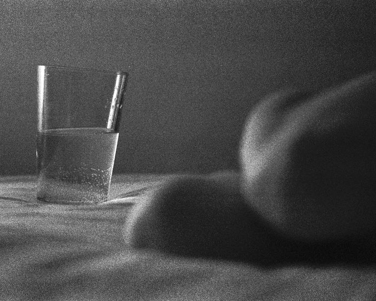 A glass of water with a pen in it sits on a textured surface next to a blurred form.