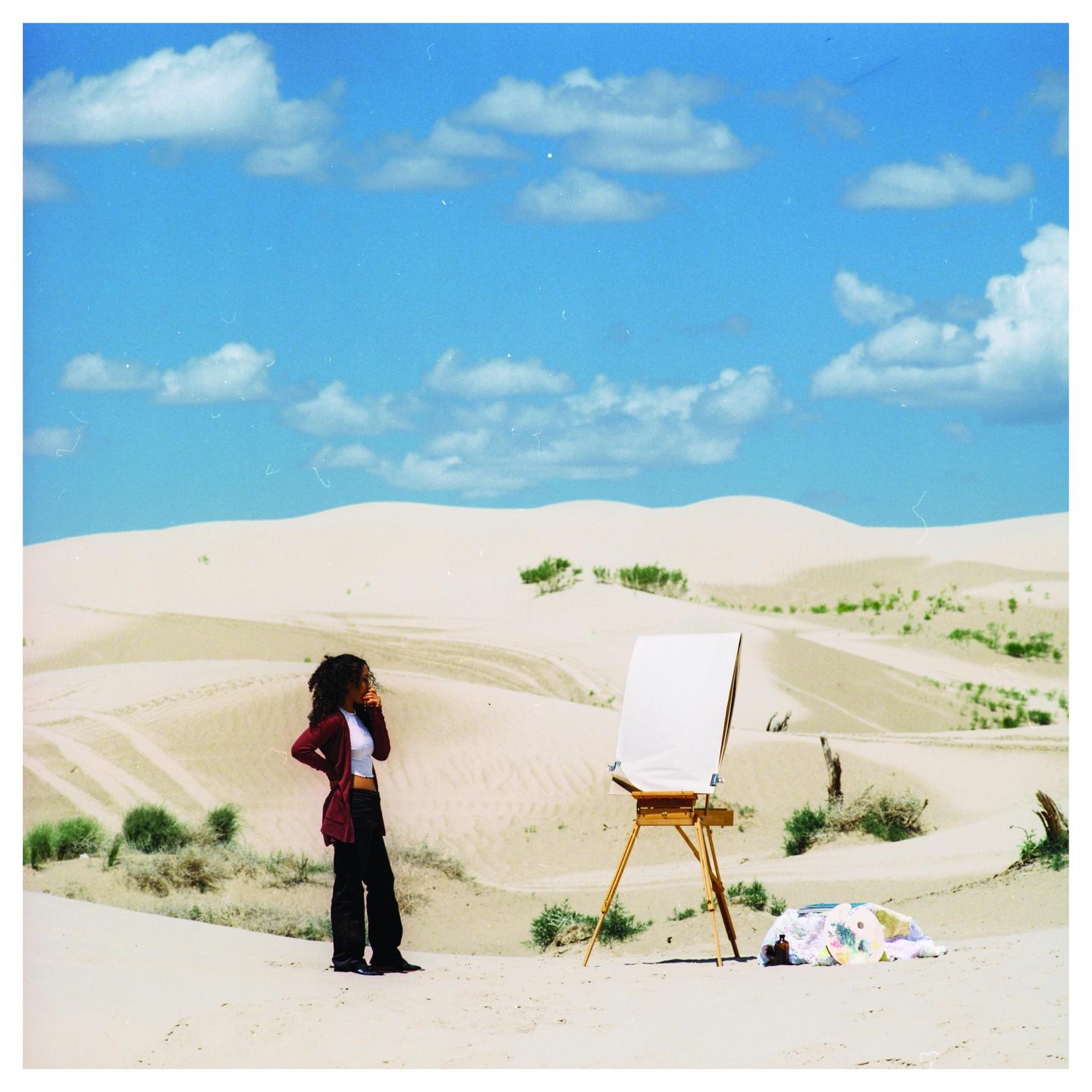 A woman stands next to an easel with a blank canvas in a desert landscape under a blue sky with clouds.