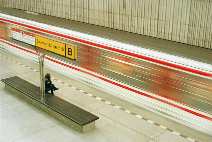 A woman sits on a bench at a subway station as a train speeds by, creating a motion blur effect.