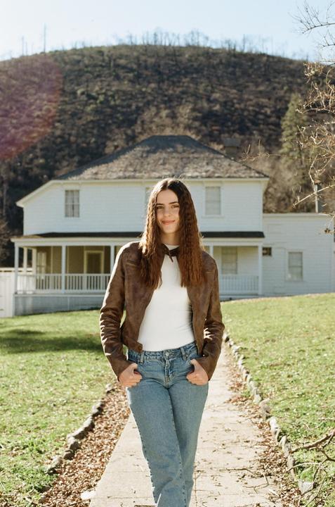 A woman stands on a path in front of a white house with a mountain in the background.