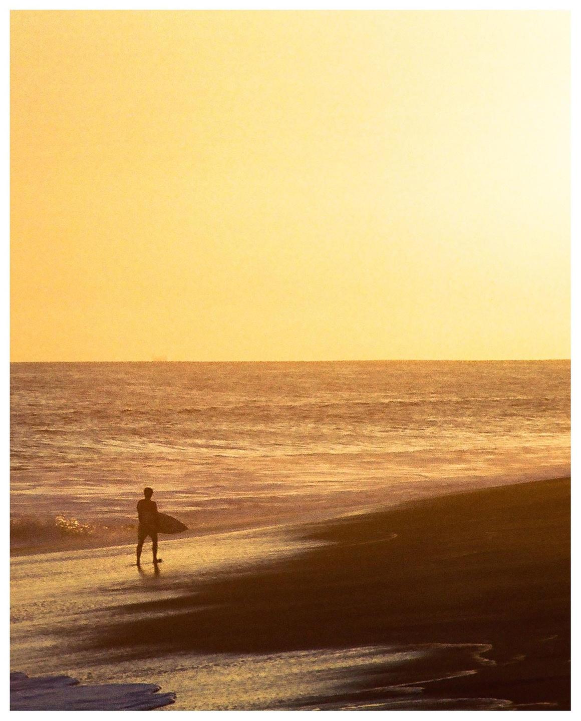 A surfer stands on the shore with a surfboard at sunset.