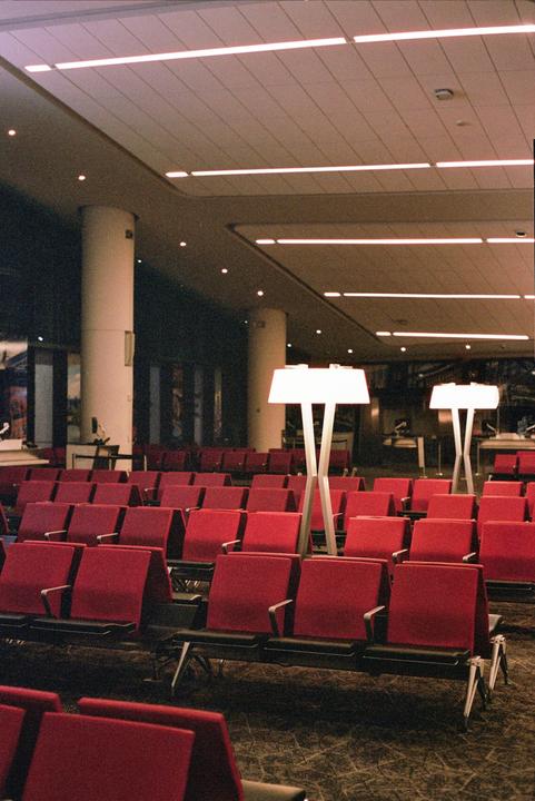 An empty airport waiting area features rows of red seats and two tall, modern lamps.