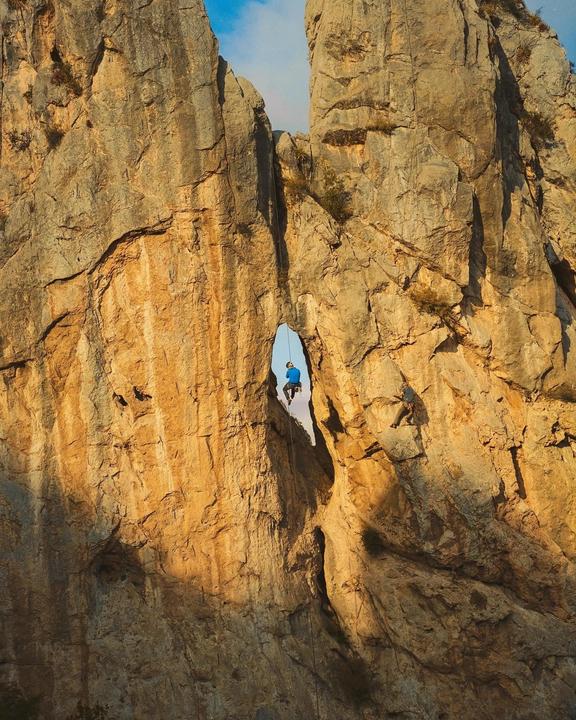 A man rappels through a natural arch in a rock face while another man climbs nearby.
