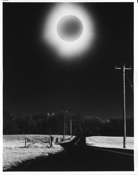 A total solar eclipse is visible above a rural road with telephone poles and a wooden fence.