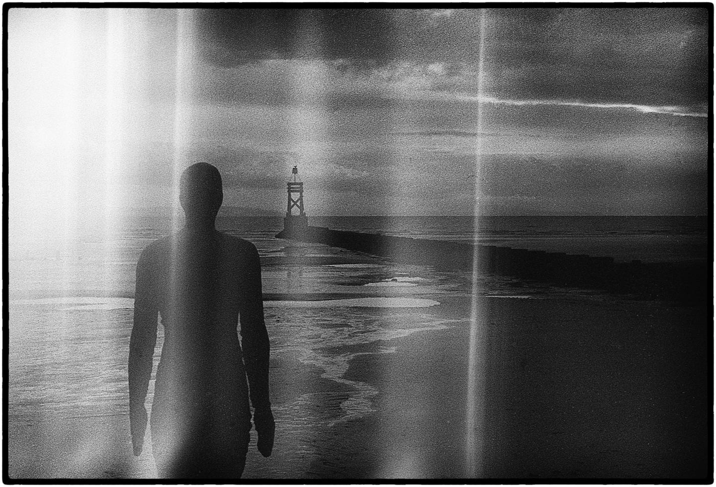 A silhouetted figure stands on a beach looking out at the ocean with a beacon in the distance.