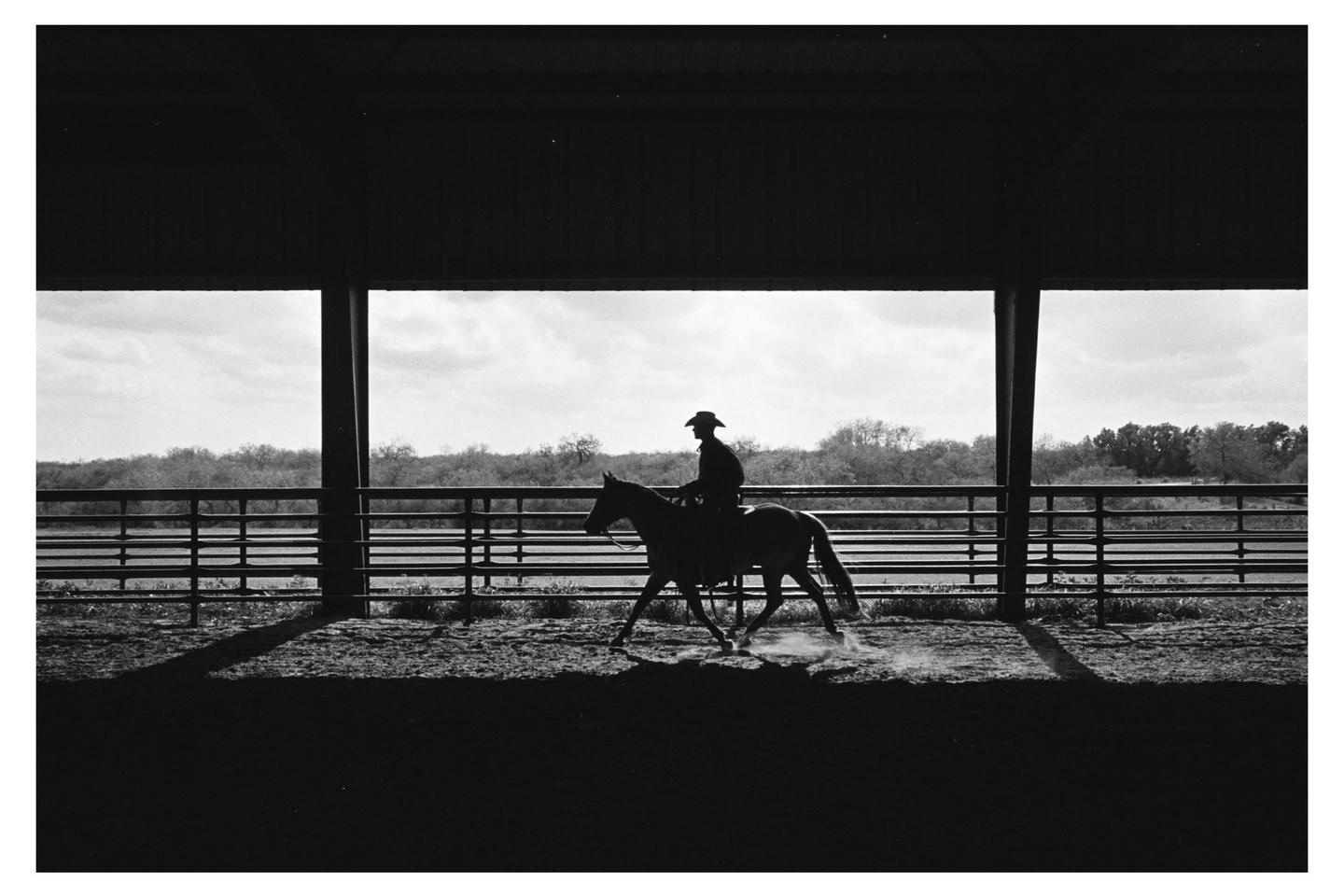 A man on horseback is silhouetted against a bright sky, riding in an arena with a fence and trees in the background.