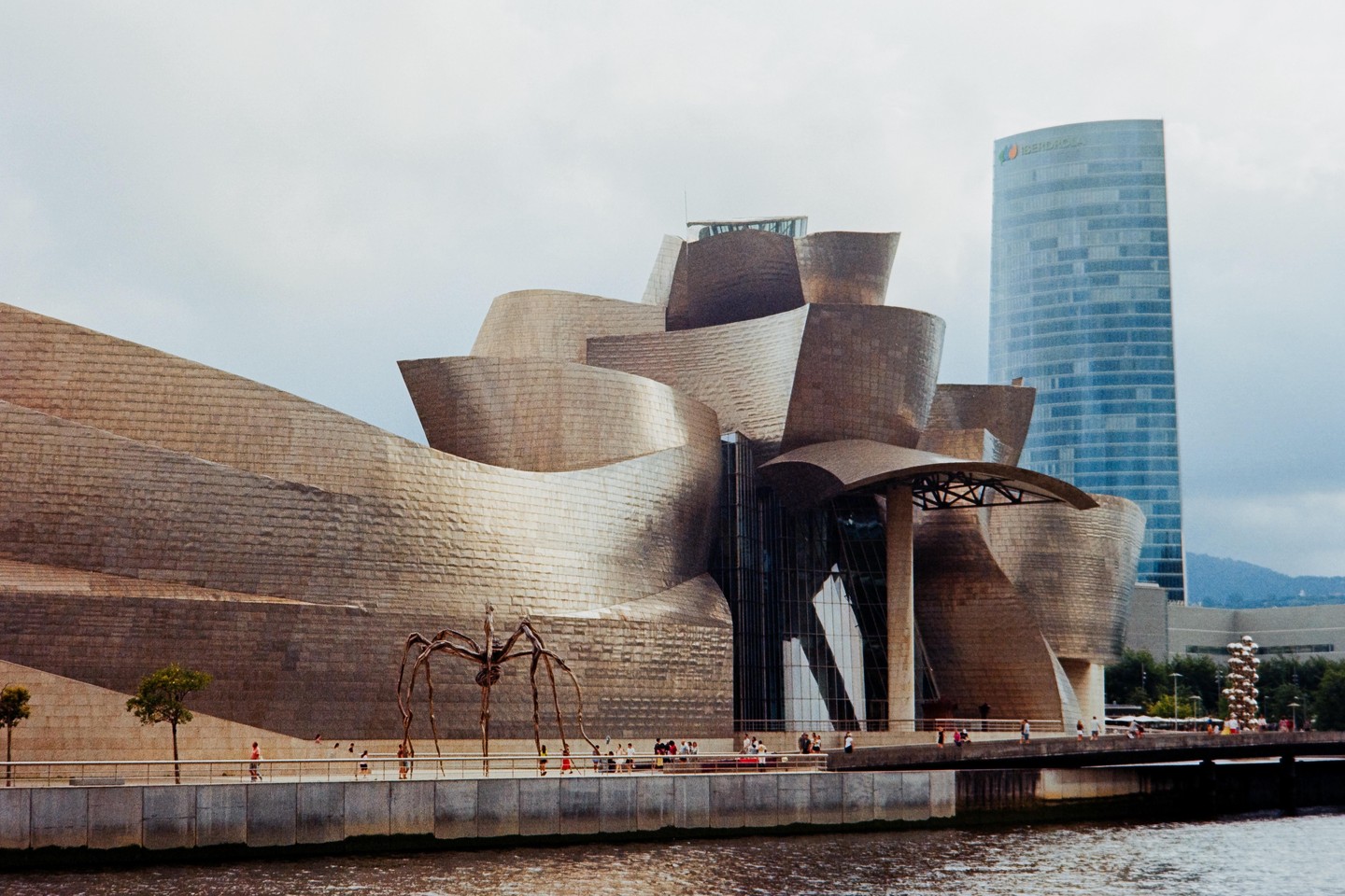 The Guggenheim Museum Bilbao with its titanium facade stands next to a river, with a tall glass building and mountains in the background.