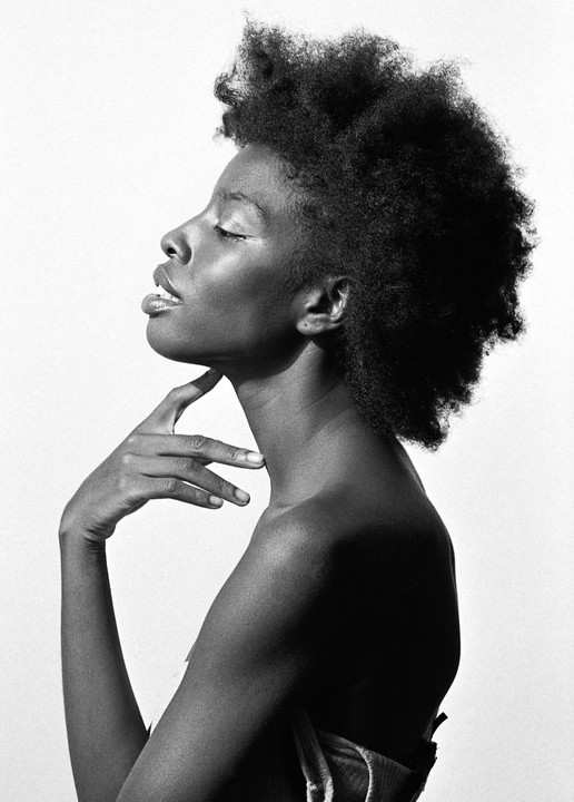 A woman with closed eyes and an afro poses in profile with her hand on her neck.