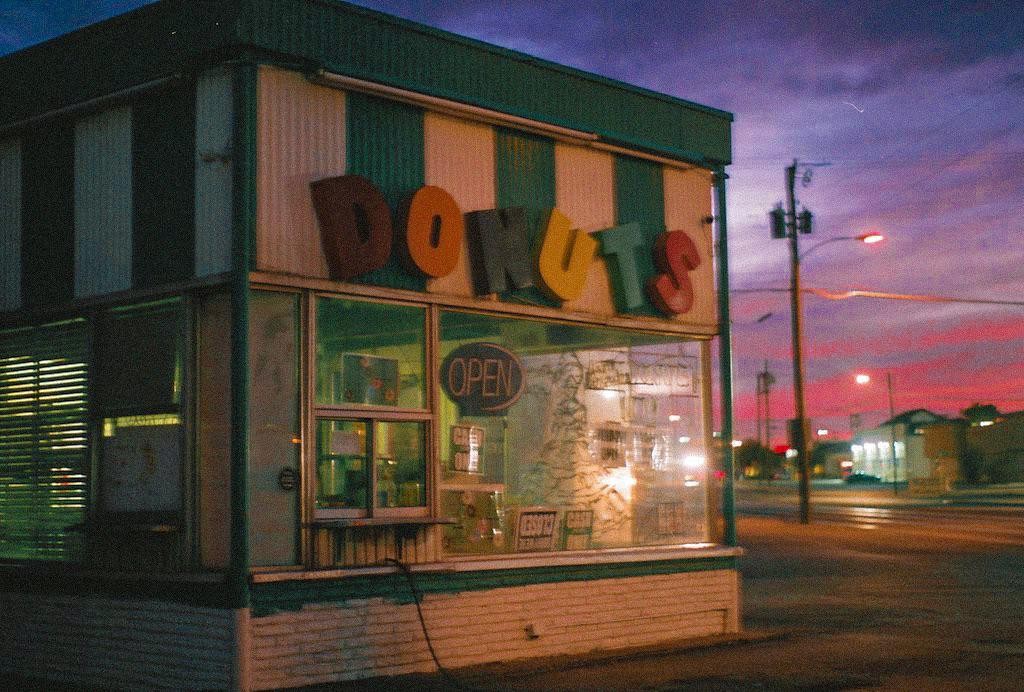 A donut shop with an "Open" sign is visible under a vibrant pink and purple sunset sky.