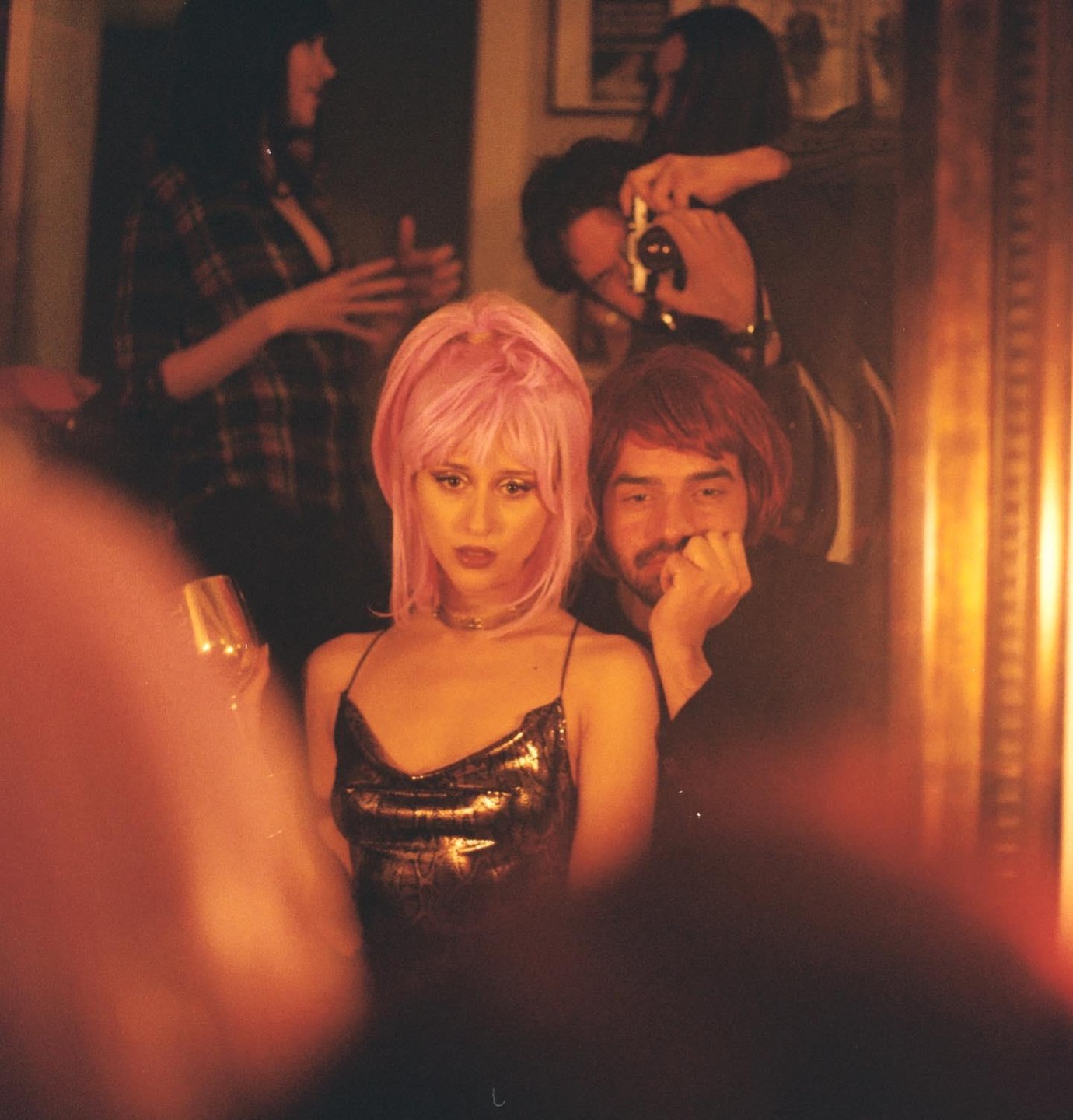 A woman with pink hair and a man with a beard are sitting together in a dimly lit room with other people in the background.