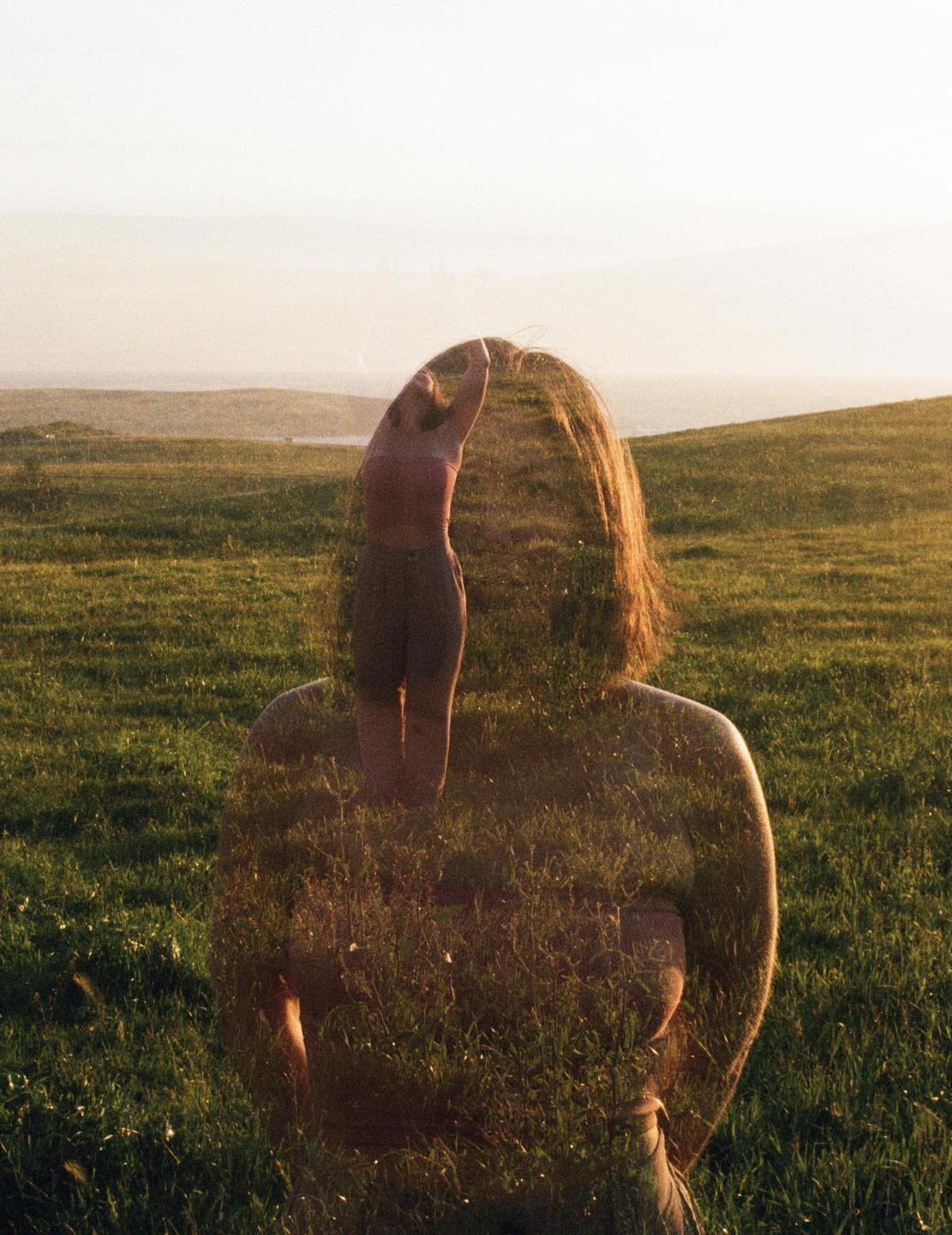 A double exposure shows a woman from behind superimposed with a woman with her arms raised in a grassy field at sunset.