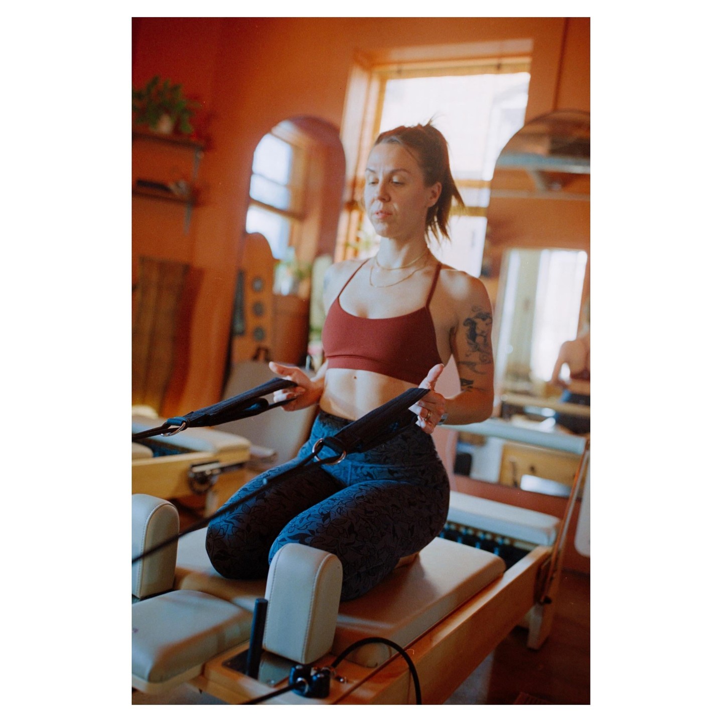 A woman kneels on a pilates reformer, pulling on the straps.