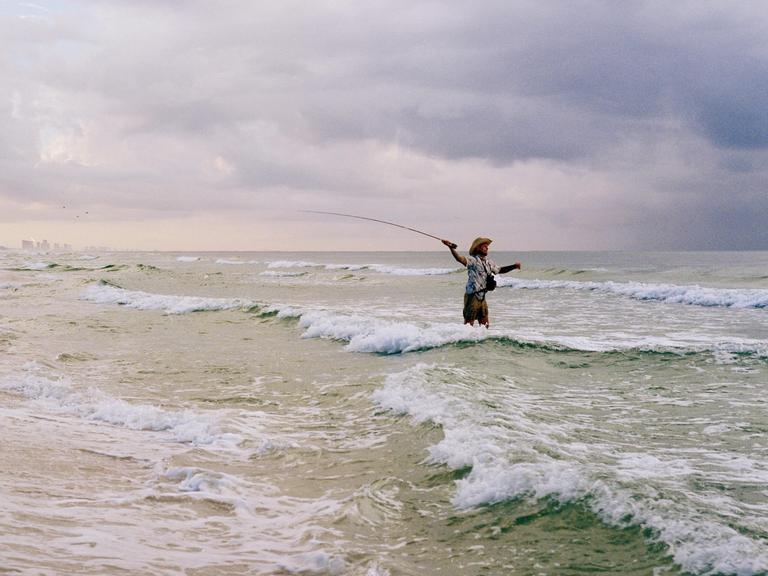A man wearing a cowboy hat stands in the ocean holding a fishing rod as waves crash around him under a cloudy sky.