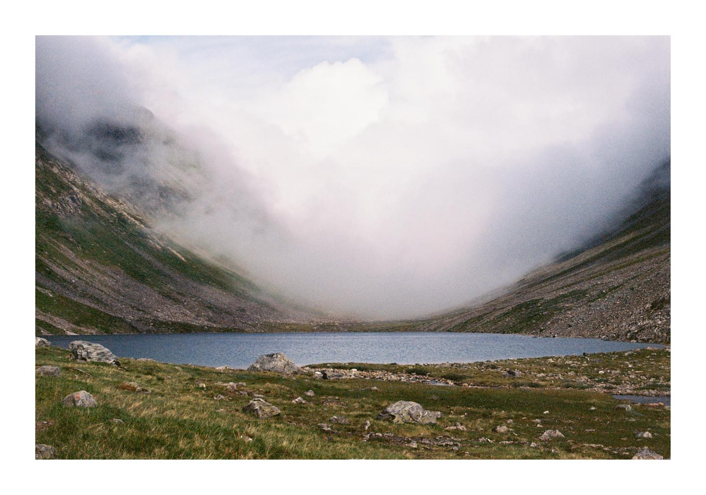 A lake is nestled between two mountains with clouds descending into the valley.
