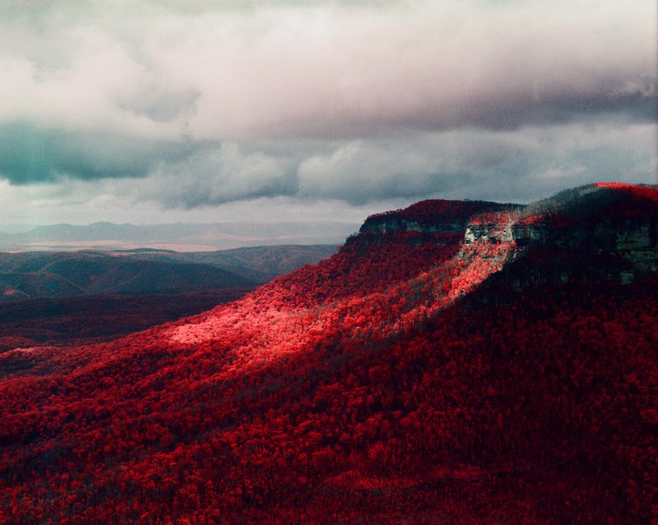A dramatic landscape shows a mountain range covered in red foliage under a cloudy sky, with a beam of light illuminating a section of the mountain.