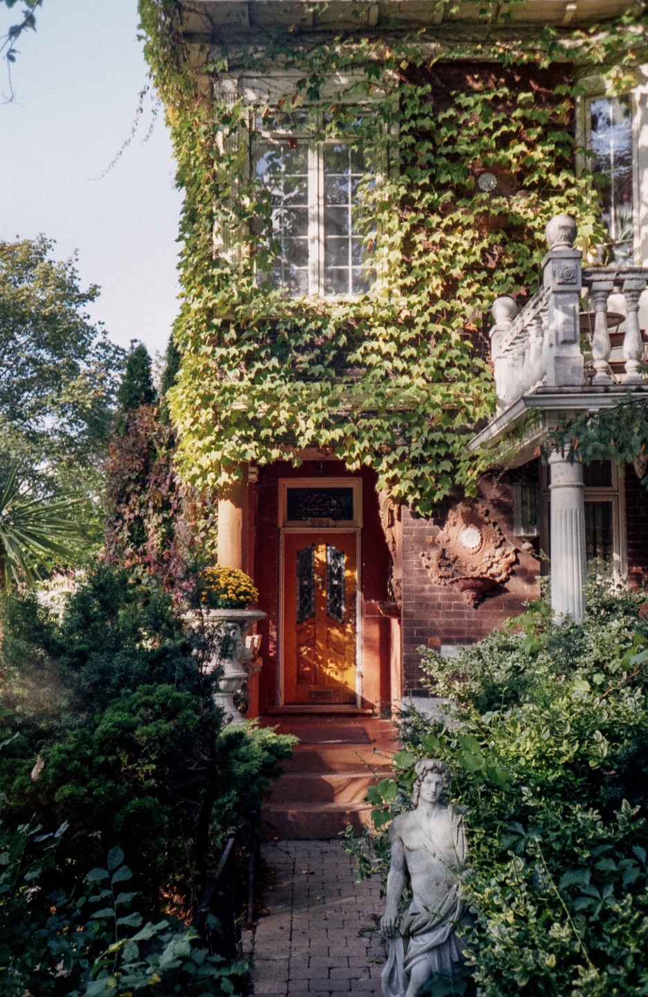 A brick house covered in ivy has a bright orange door and a statue in the garden.