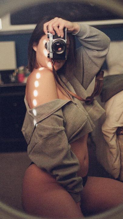A woman takes a self-portrait in a mirror, holding a camera to her face, with light spots on her shoulder.