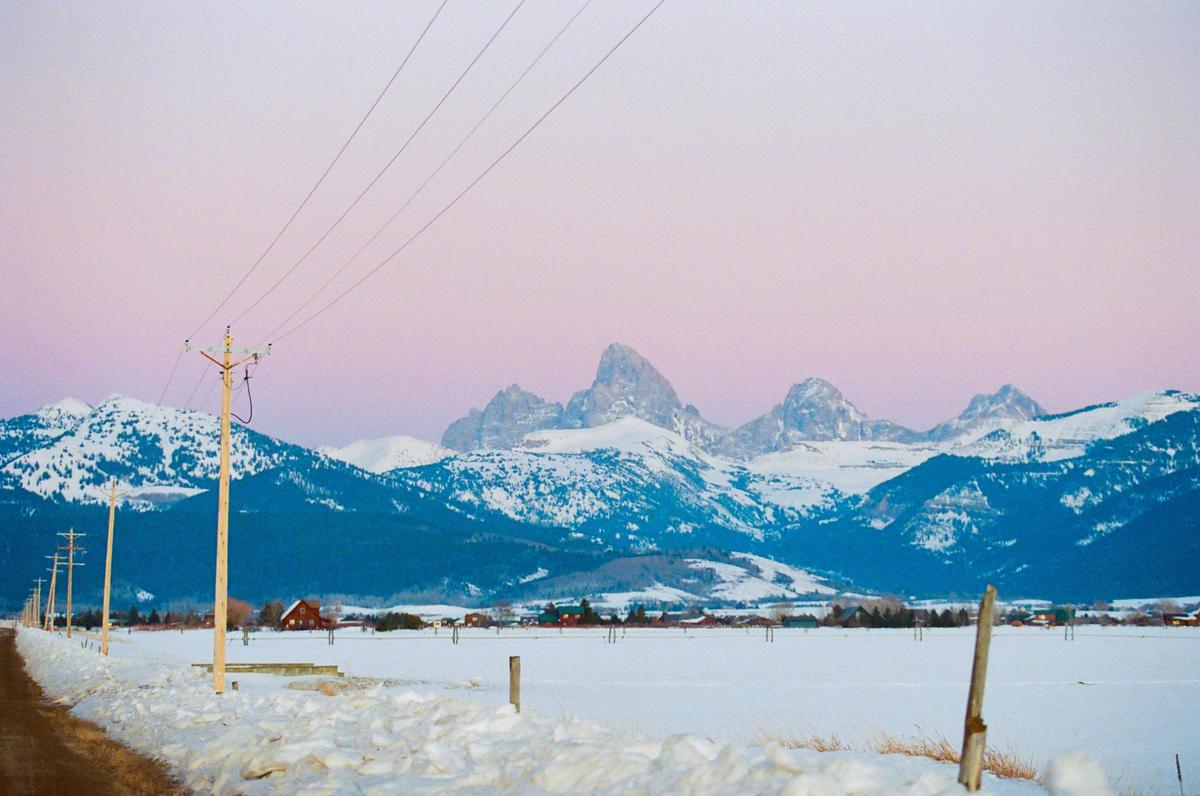 A snow-covered field and road lead towards a mountain range under a pink and purple sky.