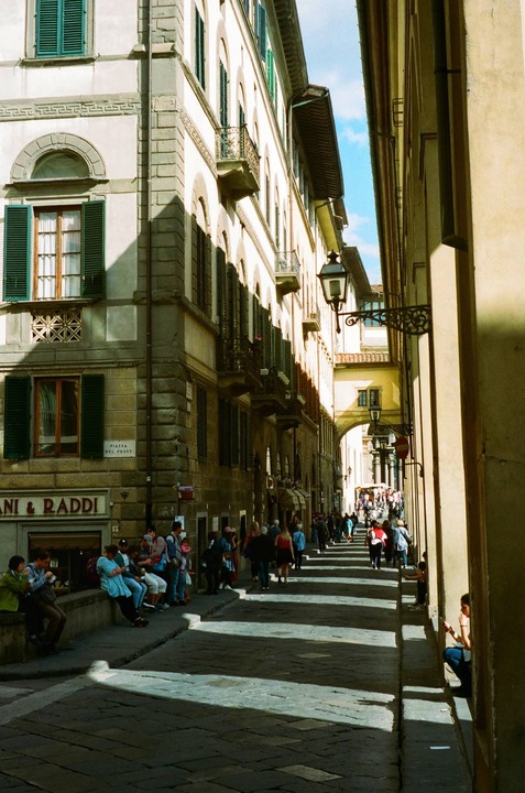 A narrow street in Florence is filled with people and dappled sunlight.