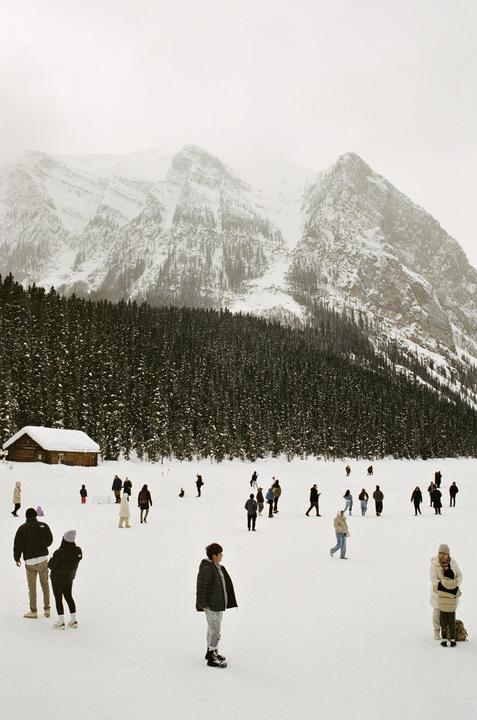 A crowd of people stands on a snow-covered field with a small cabin, a forest, and snow-capped mountains in the background.