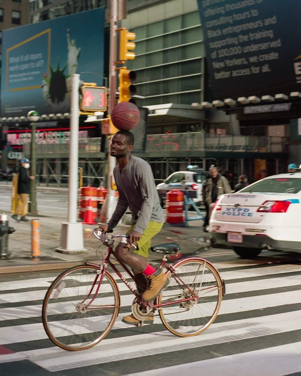 A man balances a basketball on his head while riding a bicycle across a crosswalk in a city.