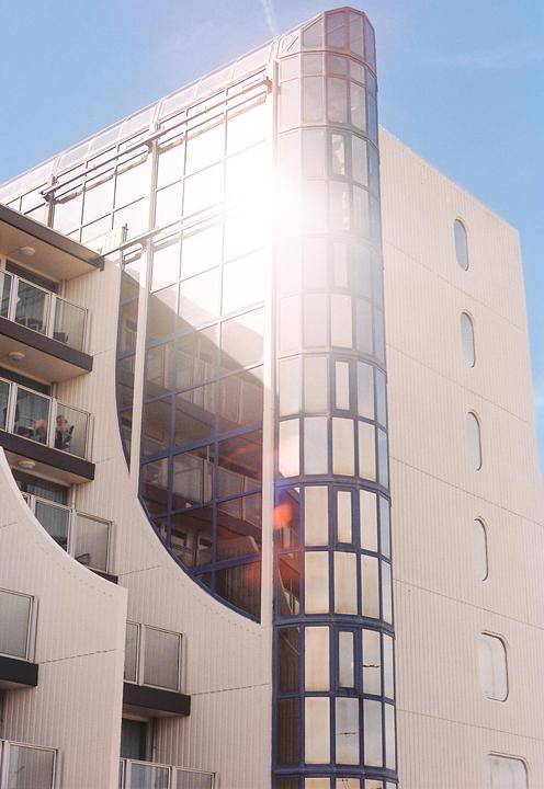 The sun reflects brightly off the glass facade of a modern building with balconies and a curved glass tower.