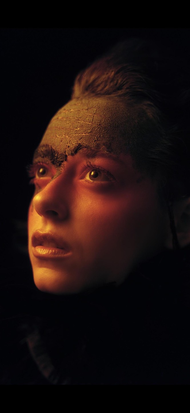 A woman with cracked mud on her forehead looks up, illuminated by warm light against a dark background.