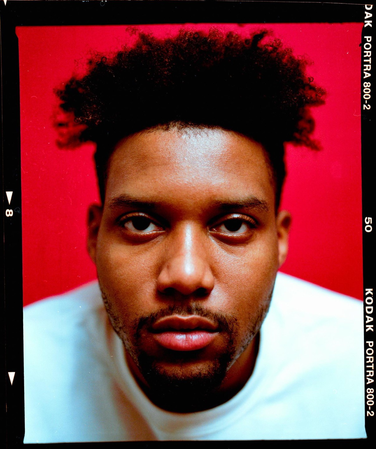 A man with dark, curly hair and a beard looks directly at the viewer against a red background.